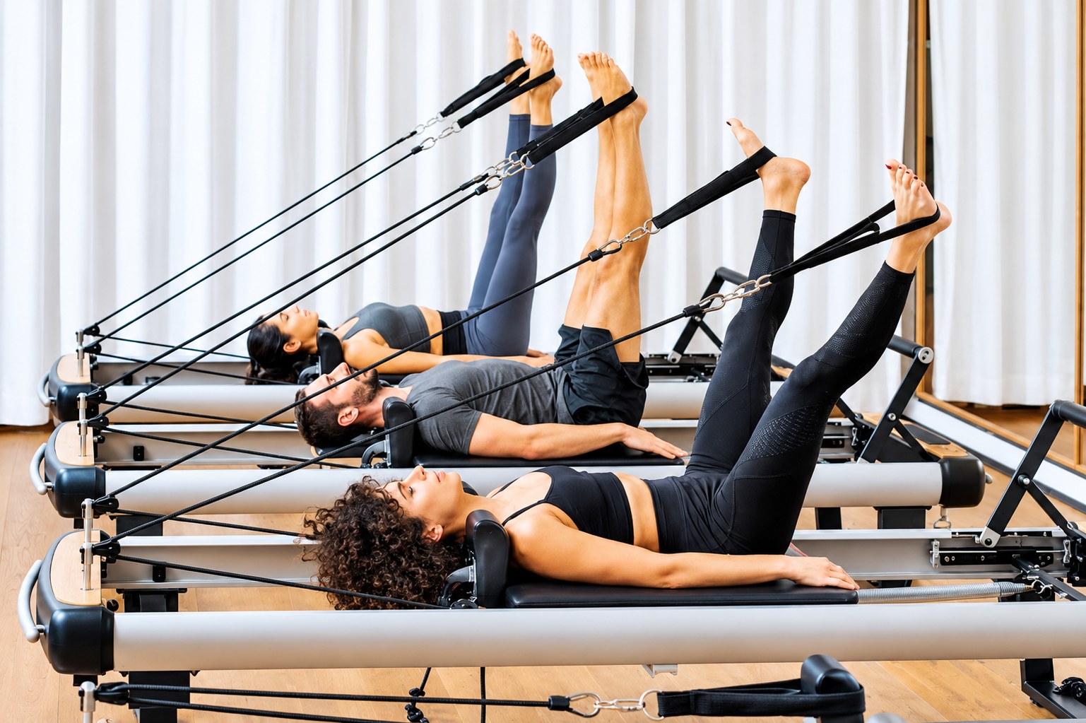 Three people practicing leg lifts on reformer machines in a bright Pilates studio